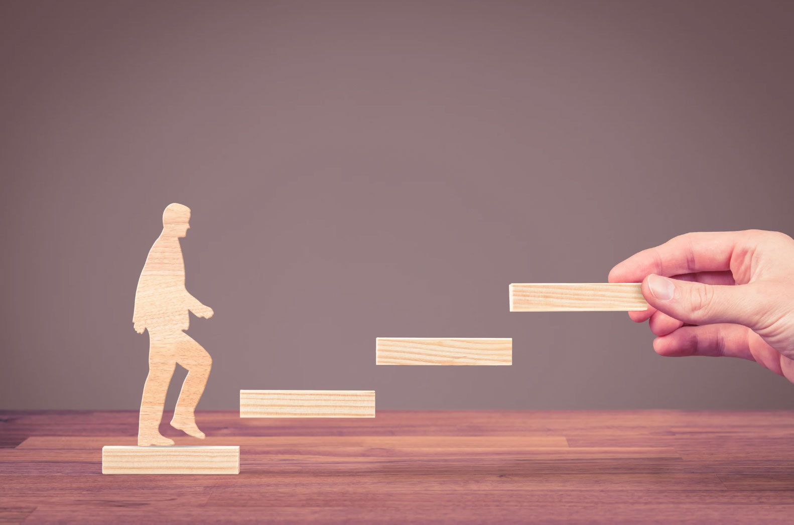 Wooden blocks arranged as ascending steps with a wooden human figure climbing upward, while a hand places the next step ahead, symbolising guidance, career progression, support, growth, and achieving future goals.