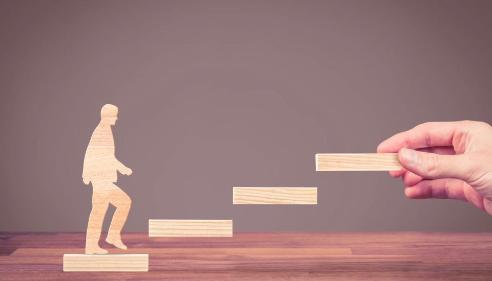 Wooden blocks arranged as ascending steps with a wooden human figure climbing upward, while a hand places the next step ahead, symbolising guidance, career progression, support, growth, and achieving future goals.