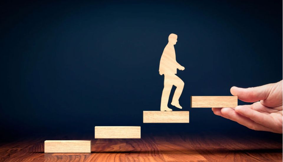 Wooden blocks arranged as ascending steps with a wooden human figure climbing upward, while a hand places the next step ahead, symbolising guidance, career progression, support, growth, and achieving future goals.