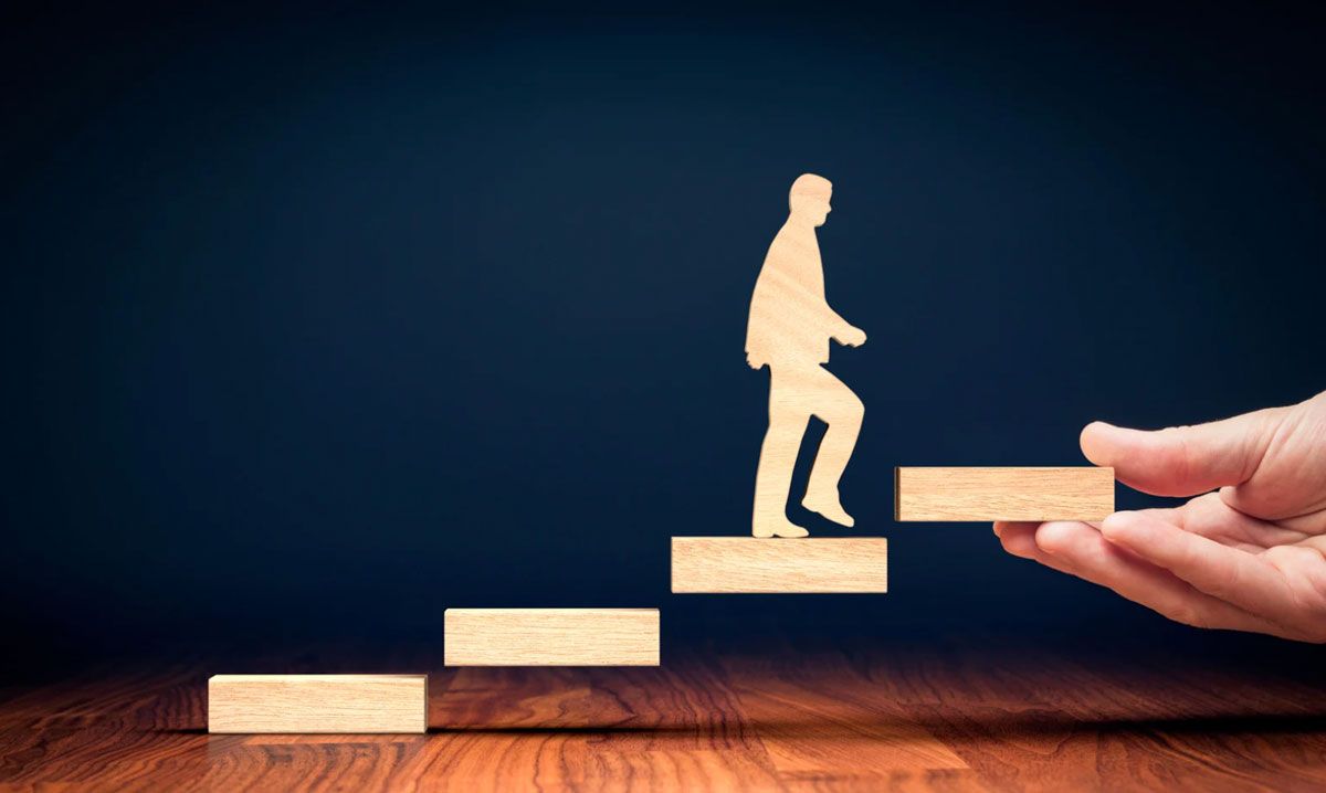 Wooden blocks arranged as ascending steps with a wooden human figure climbing upward, while a hand places the next step ahead, symbolising guidance, career progression, support, growth, and achieving future goals.
