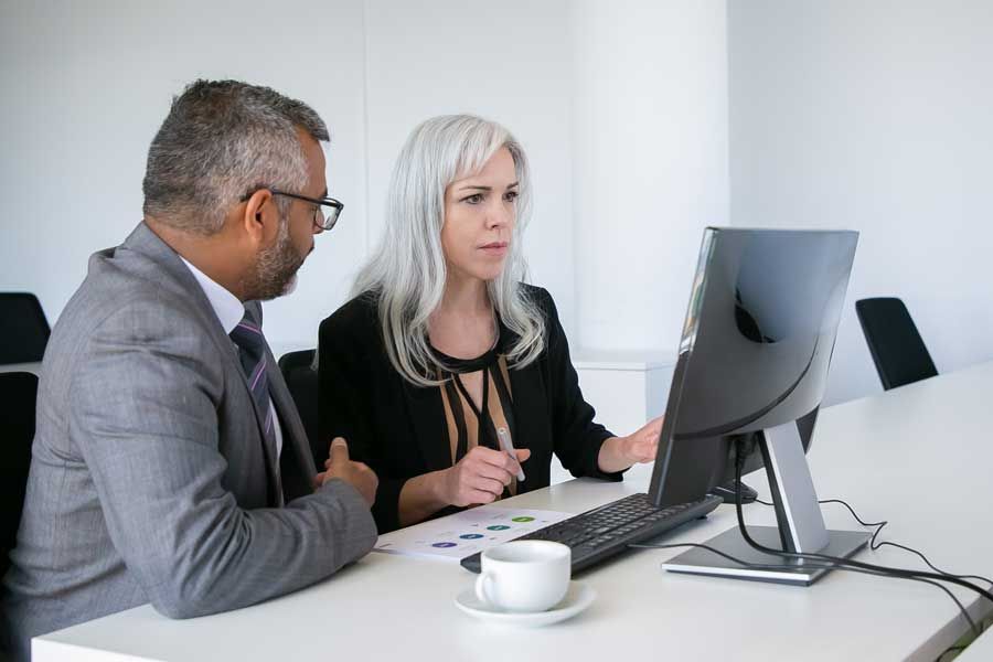 A man and woman in business attire are focused on a computer screen, collaborating on a project together.