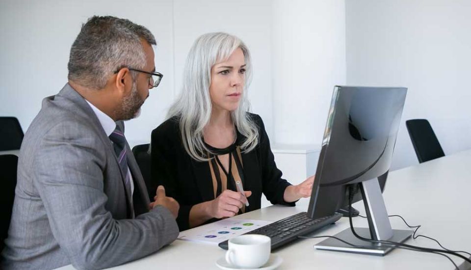A man and woman in business attire are focused on a computer screen, collaborating on a project together.